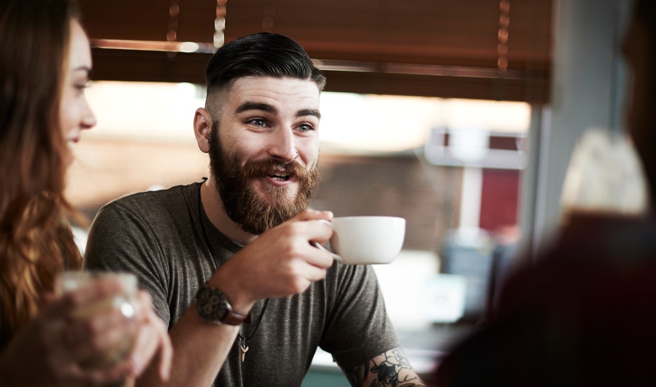 Young bearded male smiling, drinking coffee with friends